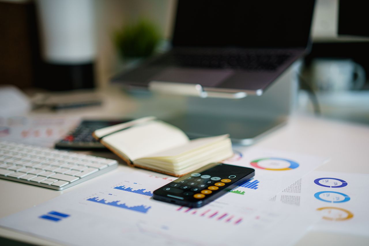 about-us-01 Modern office desk with a laptop, smartphone calculator app, and financial charts.